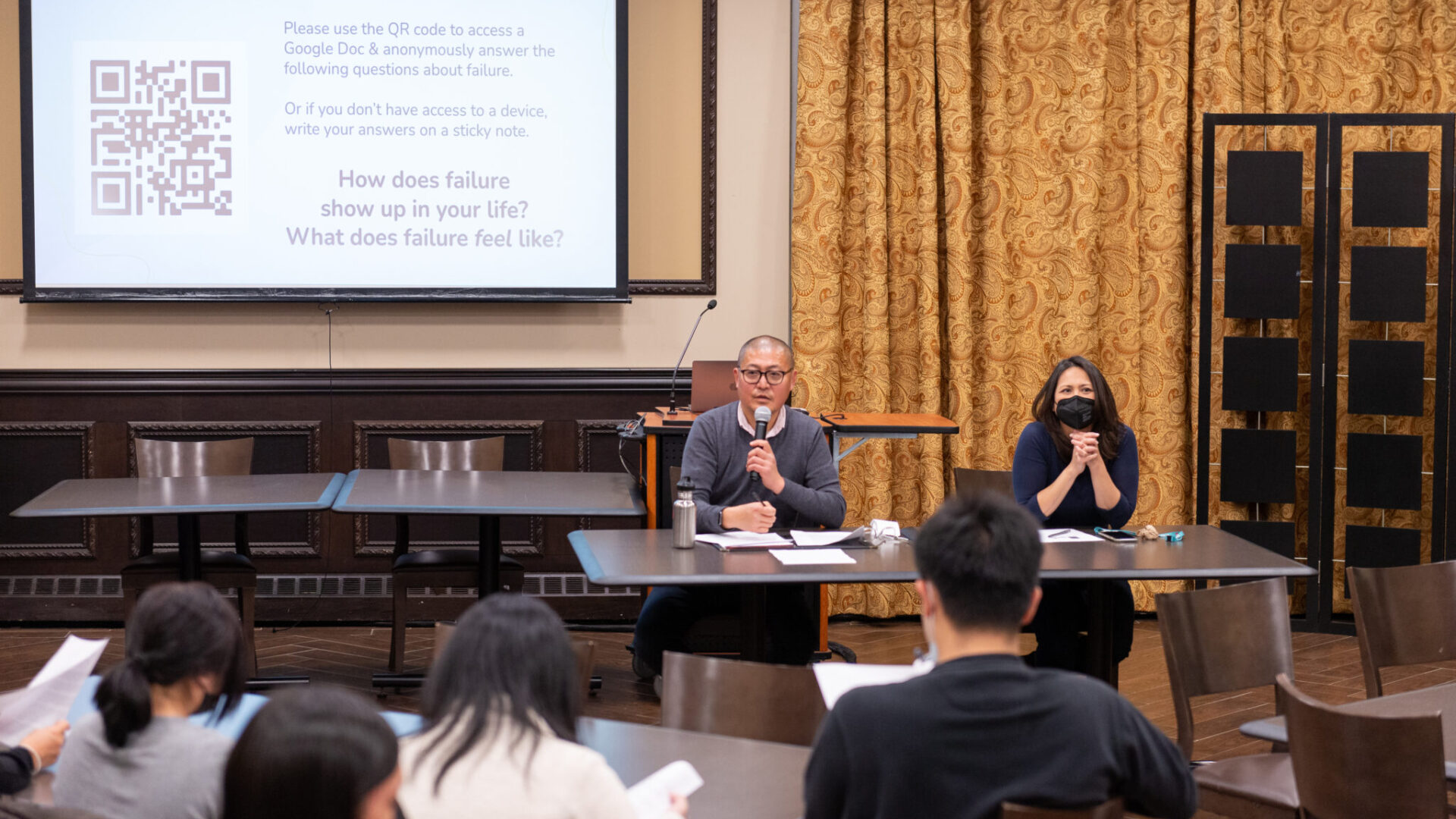 Dr. James Kyung-Jin Lee and Dr. erin Khuê Ninh lead a QCAP Mental Health Workshop, "#AsianFail: Illness and Ingratitude." Lee and Ninh are seated at a table in front of a projection screen.